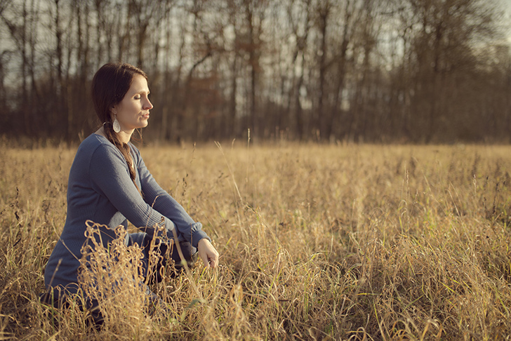 Deutschland, Bayern, Junge Frau sitzt im herbstlichen Gras einer Auenlandschaft und genießt die Sonne