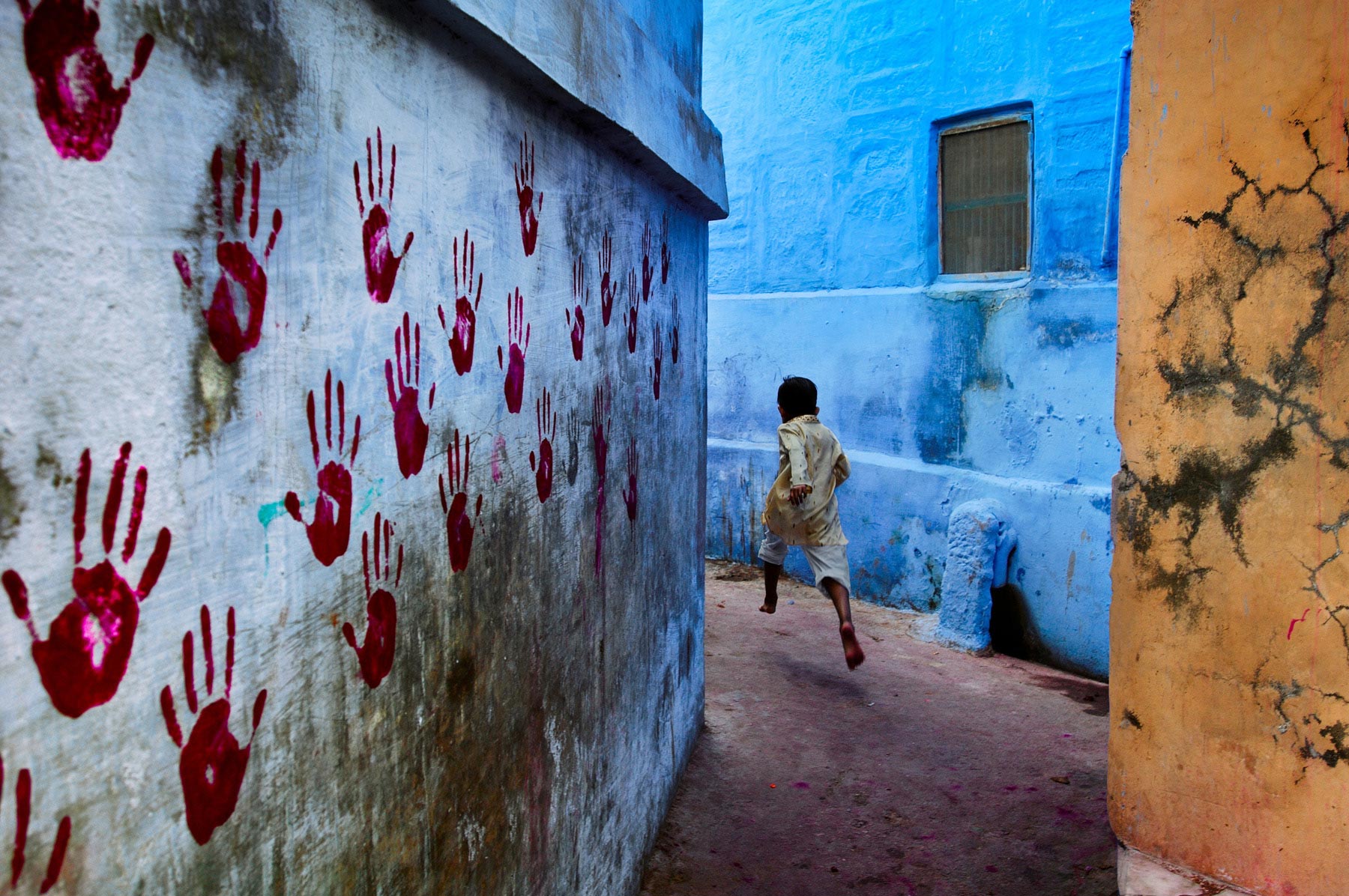 Boy in mid-flight, Jodhpur, India, 2007 At the foot of the vast Mehrangarh Fort, one can find the Blue City, a small tightly knit maze of houses located towards the north of Jodhpur. In one of the narrow alleyways a boy flees McCurry's camera. Balancing three intersecting planes of colour - one of which is covered in stark red handprints - the image pulsates with energy as a young boy dashes through the narrow alleyways. At the foot of the vast Mehrangarh Fort, one can find the Blue City, a small tightly knit maze of houses located towards the north of Jodhpur. Balancing three intersecting planes of colour - one of which is covered in stark red handprints - the image pulsates with energy as a young boy dashes through the narrow alleyways. Phaidon, The Unguarded Moment, Iconic Photographs, IP page 6, final print_poster